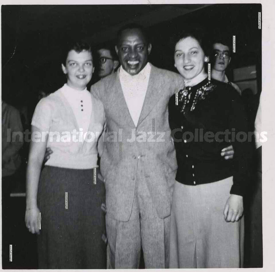 3 1/2 x 3 1/2 inch photograph. Lionel Hampton with two women, [in Canada]. Handritten on the back of the photograph: Josi Bonomo (right), (Montreal); Joan Miller (left), (Montreal)