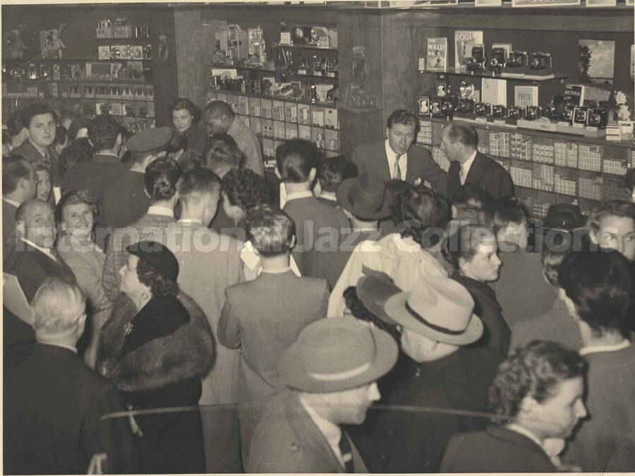 7 x 9 inch photograph. Lionel Hampton autographing for fans in a store [abroad?]. On display in this room are photographic cameras and other articles. Seen on the background are the words: Leica; Rollei