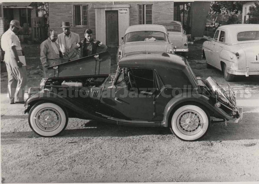3 1/4 x 4 1/2 inch photograph. Gladys Hampton standing beside a car with some members of the Lionel Hampton's orchestra, which includes guitarist Billy Mackel