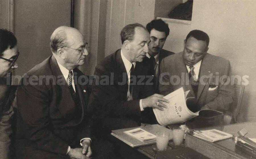 3 3/8 x 5 3/8 inch photograph in the format of postcard. Lionel Hampton looks at a publication with four unidentified men [in Israel]. Printed on the cover of the publication: Tnuva