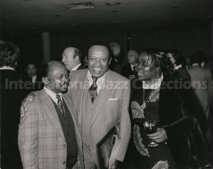8 x 10 inch photograph. Lionel Hampton with unidentified man and woman, holding a plaque from the United States Mission that appointed him as Ambassador of Music to the United Nations. Handwritten on the back of the photograph: Ambassador from Liberia