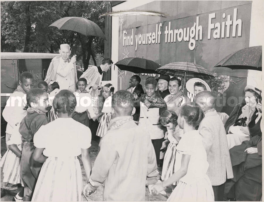 8 x 10 inch photograph. Lionel Hampton sits outside with unidentified persons, including representatives of the catholic church, in front of a wall that reads: Find yourself through Faith. The persons are observing the children holding hands in a circle