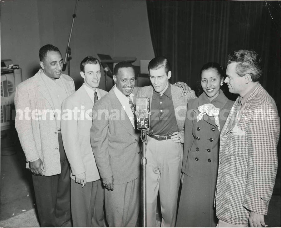 8 x 10 inch photograph. Lionel Hampton stands with Doug Duke on his right and four unidentified persons around an NBC microphone