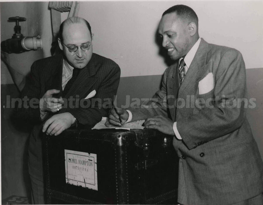 8 x 10 inch photograph. Lionel Hampton goes over sheet music with unidentified man. They are leaning on a box that reads: Lionel Hampton orchestra