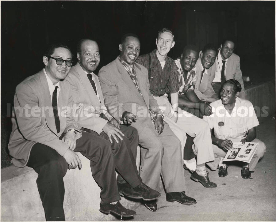 8 x 10 inch photograph. Lionel Hampton sits on a wall with some members of his band, including guitarist Billy Mackel and [drummer Curley Hamner]