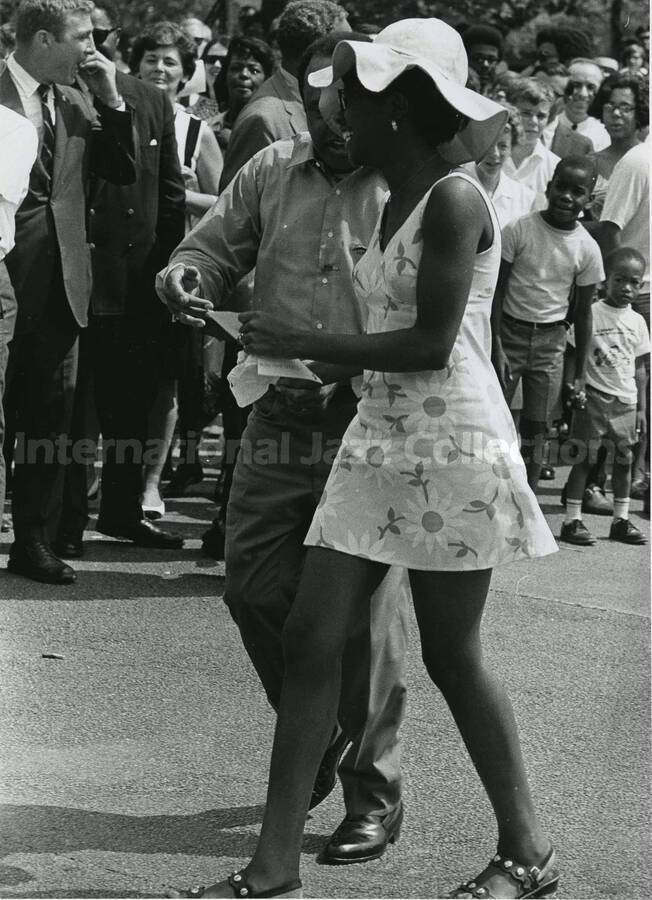 10 x 8 inch photograph. Lionel Hampton walking down a street with unidentified woman, a crowd of onlookers is in the background