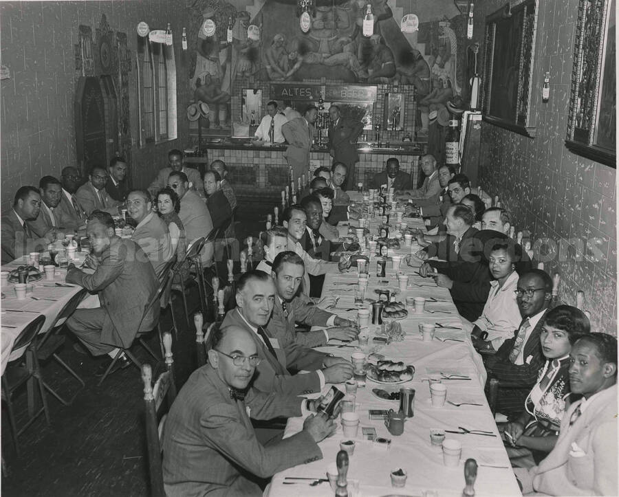 8 x 10 inch photograph. Lionel Hampton dining with unidentified persons at the tap room of the Altes Brewery, in Logan Heights, San Diego, CA. Seen on the back and lateral walls of the room are the Mexican murals painted by Jose Moya del Pino, in 1934, for the former Aztec Brewery