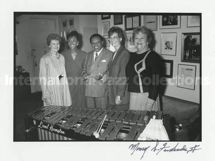 8 x 10 inch photograph. Lionel Hampton standing behind his vibraphone with four unidentified women, in front of a wall displaying plaques and certificates, in his apartment