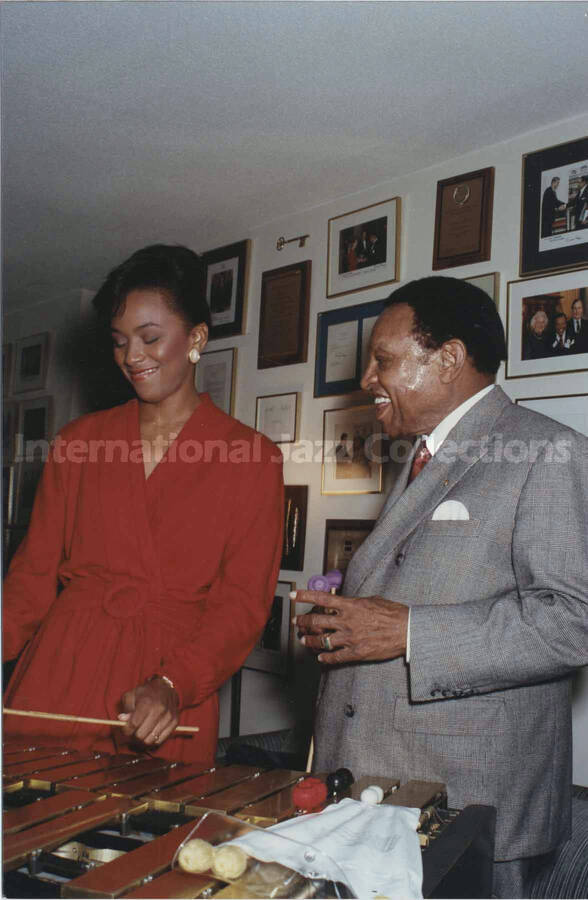6 x 4 inch photograph. Lionel Hampton playing the vibraphone with an unidentified woman, in front of a wall displaying plaques and certificates, in his apartment