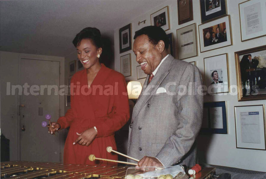 4 x 6 inch photograph. Lionel Hampton playing the vibraphone with an unidentified woman, in front of a wall displaying plaques and certificates, in his apartment. Also an 8 3/4 x 13 1/4 copy of the same photograph mounted on cardboard, signed by the photographer (LH.III.2470)