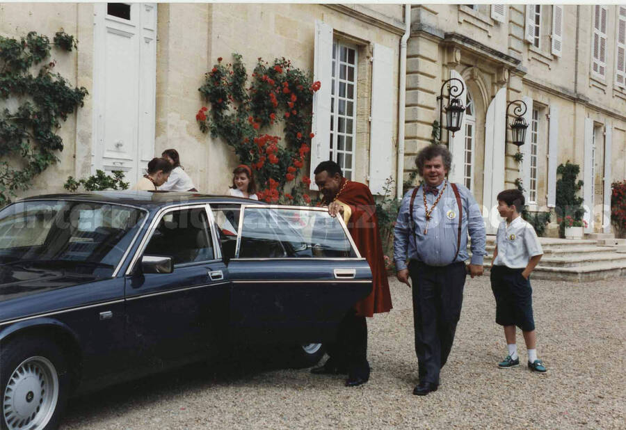 4 x 6 inch photograph. Lionel Hampton gets into a car wearing a red cloak, on the occasion of a ceremony related to wine, [in France]. An unidentified man stands by wearing a key hanging from a red and gold string around his neck