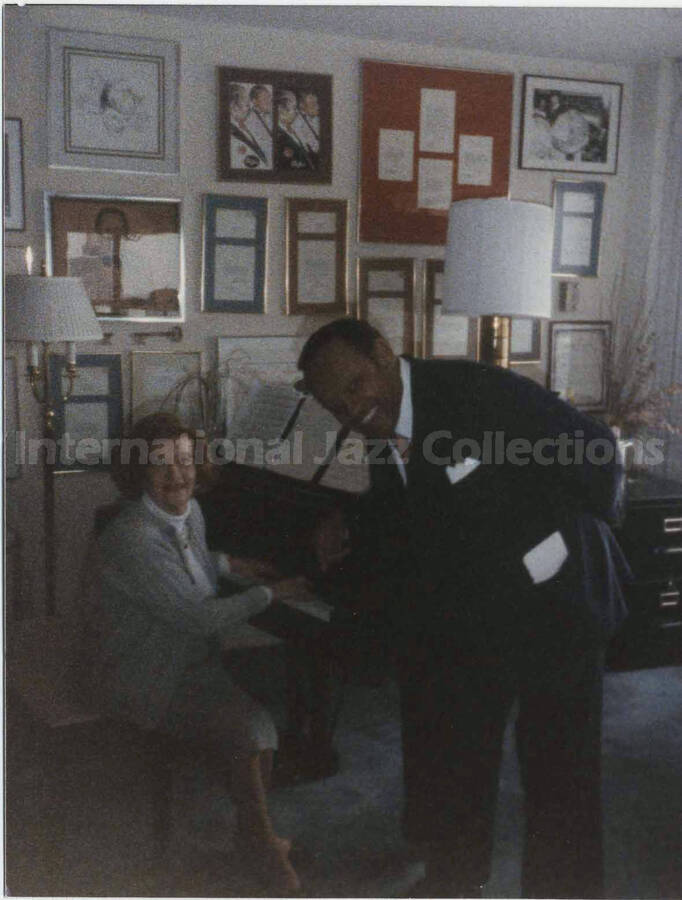 5 x 4 inch photograph. Lionel Hampton with unidentified woman sitting at the piano, in front of a wall displaying plaques and certificates, in his apartment
