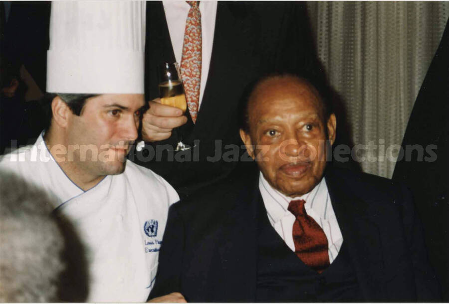 4 x 6 inch photograph. Lionel Hampton with a chef at a reception. The chef's jacket includes the emblem of the United Nations