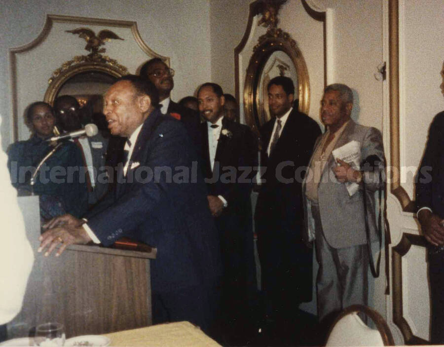 4 x 5 inch photograph. Lionel Hampton speaks at a podium observed by unidentified musicians at a reception