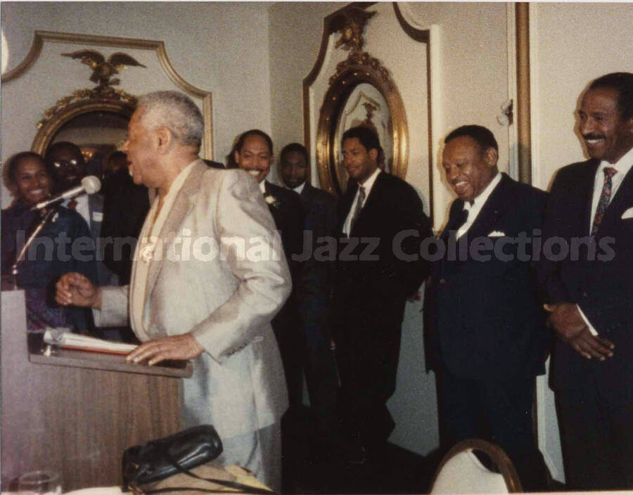 4 x 5 inch photograph. Dizzy Gillespie speaks at a podium observed by unidentified musicians at a reception