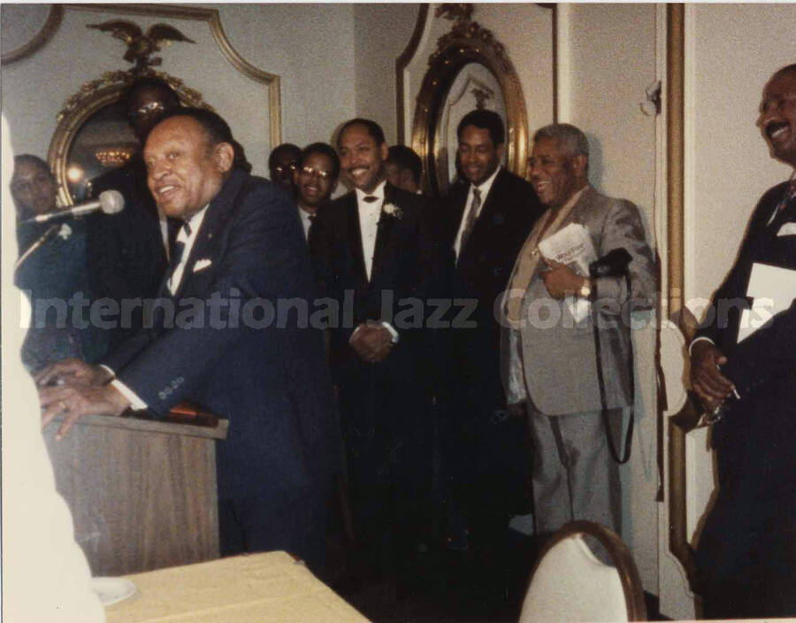 4 x 5 inch photograph. Lionel Hampton speaks at a podium observed by unidentified musicians at a reception