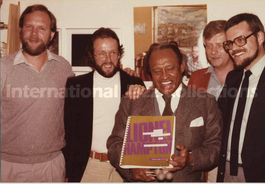 3 1/2 x 5 inch photograph. Lionel Hampton poses holding a music book with Jean Claude Forestier and unidentified men