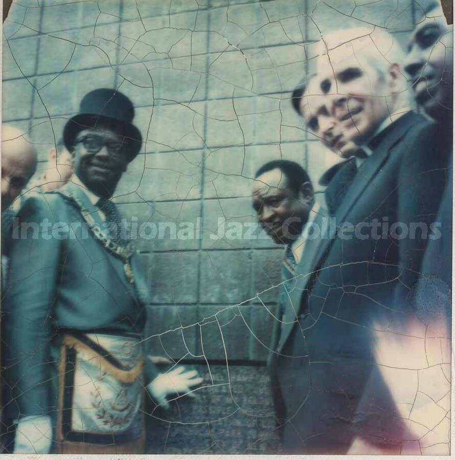 4 1/4 x 3 1/2 inch photograph. Lionel Hampton standing with unidentified member of the Medina Temple and others by a dedication plaque on a wall at the Grand Opening of Lionel Hampton Houses