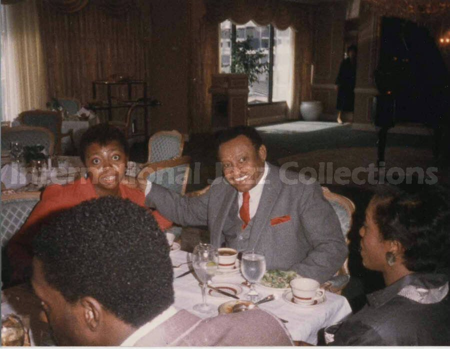 3 1/2 x 4 1/2 inch photograph. Lionel Hampton sits at a dinner table with unidentified persons