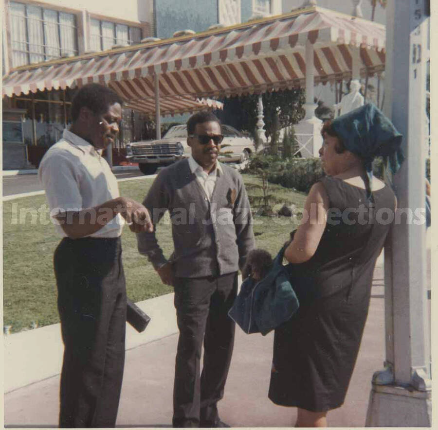 3 1/2 x 3 1/2 inch photograph. Gladys Hampton standing outside with two unidentified men. She has a dog inside her blue bag