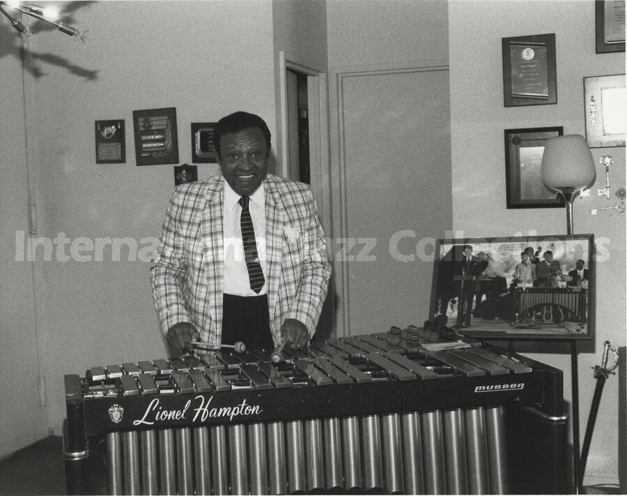 8 x 10 inch photograph. Lionel Hampton at the vibraphone in front of a wall displaying plaques and certificates, in his apartment