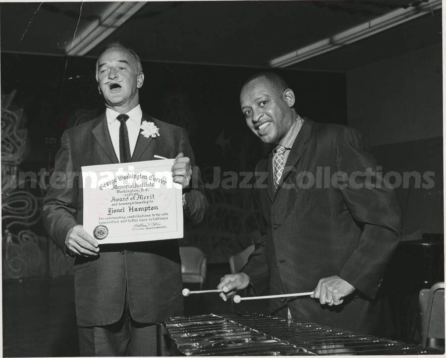 8 x 10 inch photograph. Lionel Hampton receiving a certificate of honorary fellowship from the George Washington Carver Memorial Institute, Washington D.C., from an unidentified man. The certificate is signed by William P. Tolley, Chairman Award Committee