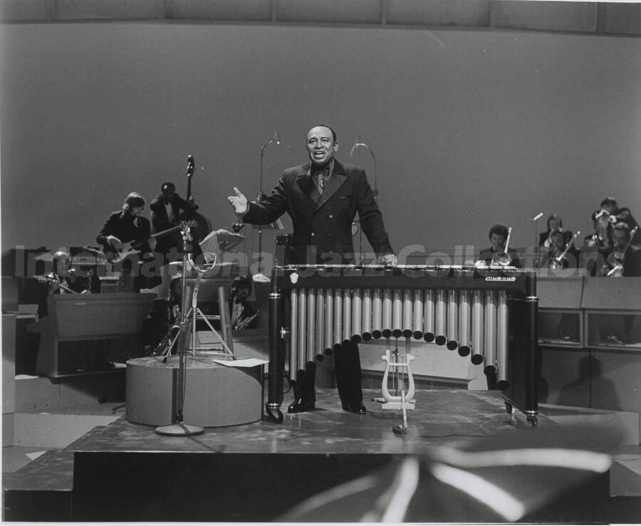 8 x 10 inch photograph. Lionel Hampton standing behind his vibraphone with band in the background, including guitarist Billy Mackel