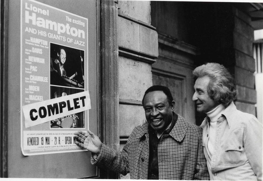 8 x 10 inch photograph. Lionel Hampton and Bill Titone looking at a poster announcing Lionel Hampton and his Giants of Jazz: Wild Bill Davison, Joe Newman, [] Pac, Eddie Chamblee, Paul Moen, and Billy Mackel, [in France]