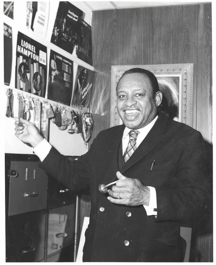10 x 8 inch photograph. Lionel Hampton, in his apartment, next to a board on which hangs his collection of hotel keys. Record albums are also visible