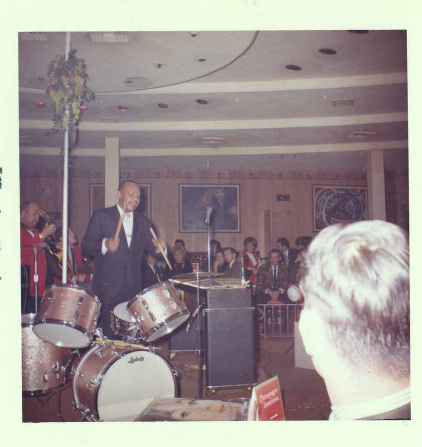 3 1/2 x 3 1/2 inch photograph. Lionel Hampton on drums with band [at Al Hirt's New Orleans Bourbon street nightclub]. Cards on the tables read: Champagne Luncheon