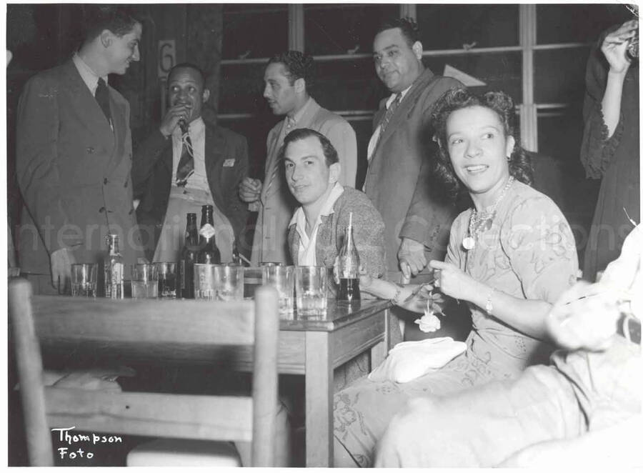 5 1/2 x 7 1/2 inch photograph. Gladys Hampton drinking Coca-Cola in a club with Lionel Hampton and unidentified men. Seen on the table is a bottle of Grand Prize beer. Handwritten on the back of the photograph: Den Bar; D & B Hotel; Hack Berry & Center