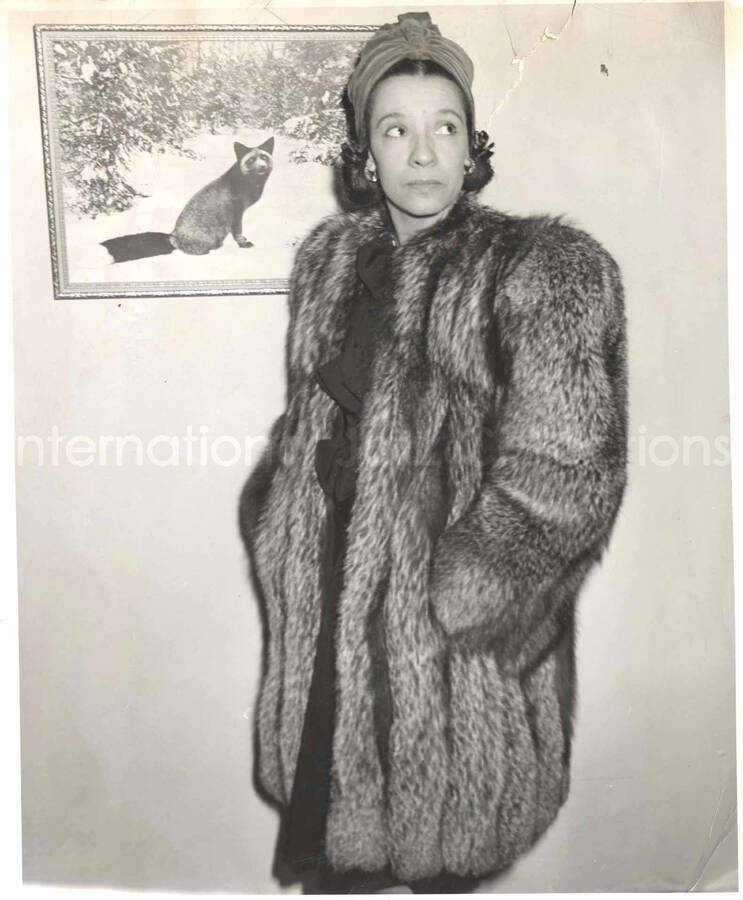 10 x 8 inch photograph. Gladys Hampton in a fox coat poses in front of a picture of a fox on a wall. Handwritten on the back of the photograph: Mrs. Lionel Hampton silver fox coat made to order by J. T. Vidal