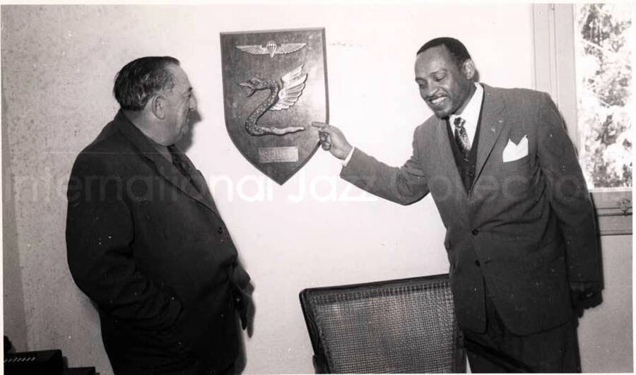 5 x 8 inch photograph. Gladys and Lionel Hampton with band in Israel. Lionel Hampton is pointing to a plaque on the wall that shows the Paratroopers Brigade Insignia