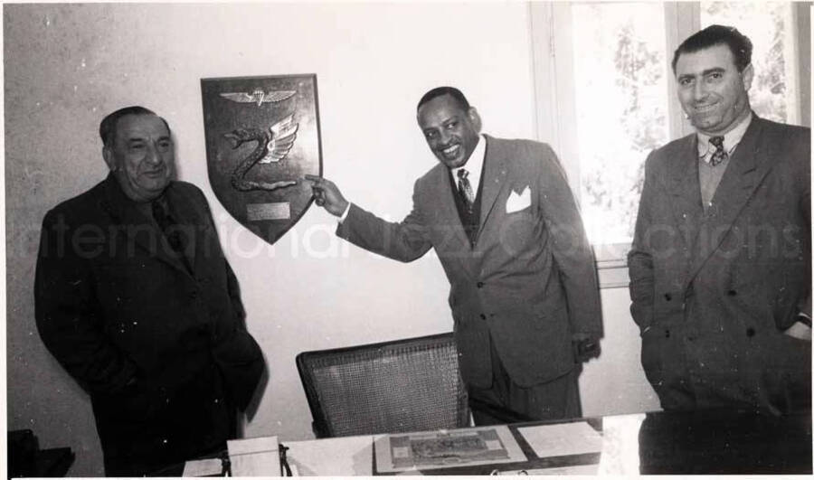 5 x 8 inch photograph. Gladys and Lionel Hampton with band in Israel. Lionel Hampton is pointing to a plaque on the wall that shows the Paratroopers Brigade Insignia