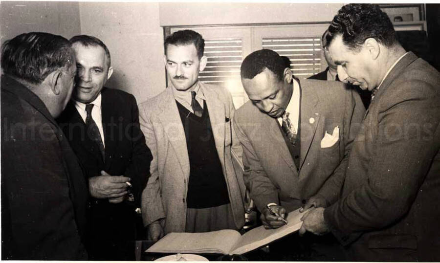 5 x 8 inch photograph. Gladys and Lionel Hampton with band in Israel. Lionel Hampton is signing a book with unidentified men