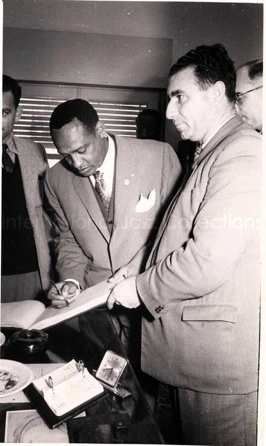 8 x 5 inch photograph. Gladys and Lionel Hampton with band in Israel. Lionel Hampton is signing a book with unidentified men