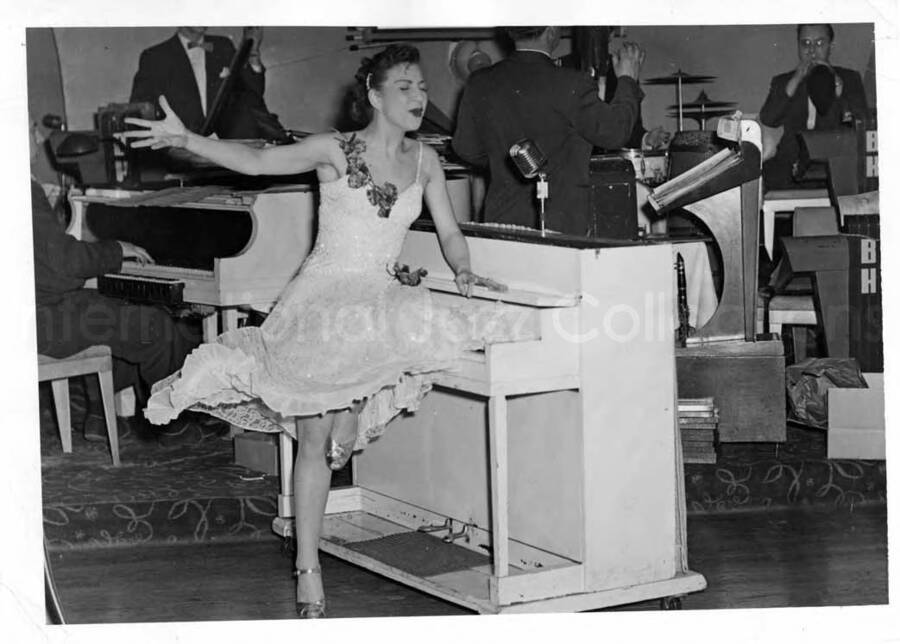 5 x 7 inch photograph. Unidentified woman dancing and singing on stage with orchestra. The musicians' stands include the initials: BH