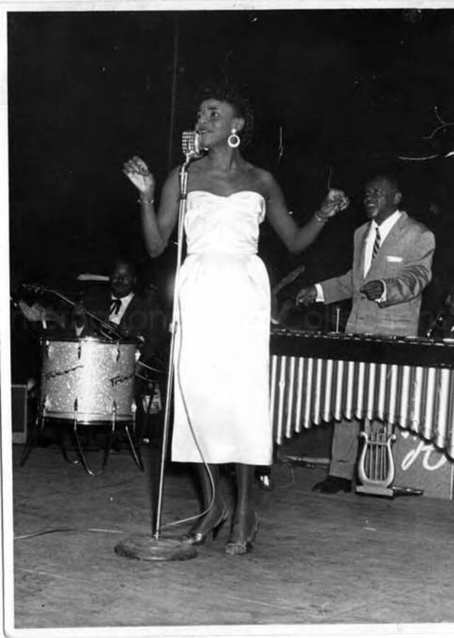 4 1/2 x 3 1/2 inch photograph. Unidentified vocalist performing with Lionel Hampton's band, which includes guitarist Billy Mackel. Lionel Hampton is at the vibraphone with a Trixon drums on his right