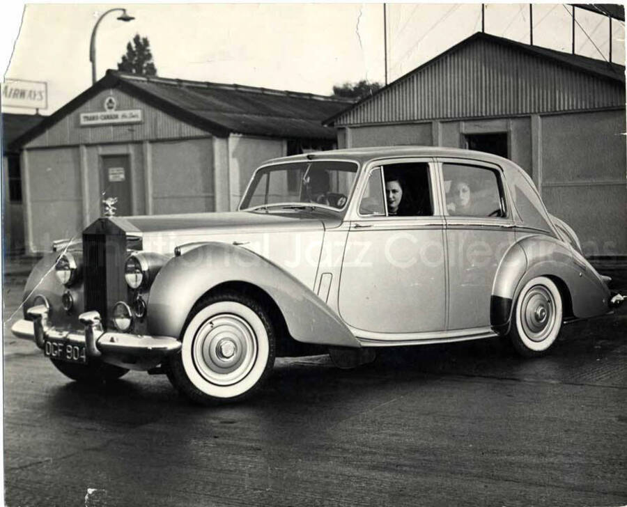 8 x 10 inch photograph. Unidentified persons inside a car parked in front of a building named the Trans-Canada. Handwritten on the back of the photograph: The Silver Pigeon meets