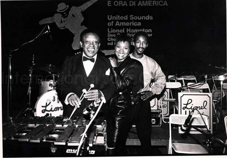 7 x 9 1/2 inch photograph. Lionel Hampton with Dee Dee Bridgewater and Chester Whitmore [in Italy]. The background of the stage reads: E ora di America; United Sounds of America