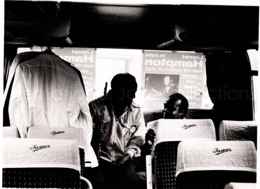 4 x 5 inch photograph. Two unidentified musicians sitting in the back of the bus. A poster of the Lionel Hampton and His Giants of Jazz is pasted on the rear window of the bus