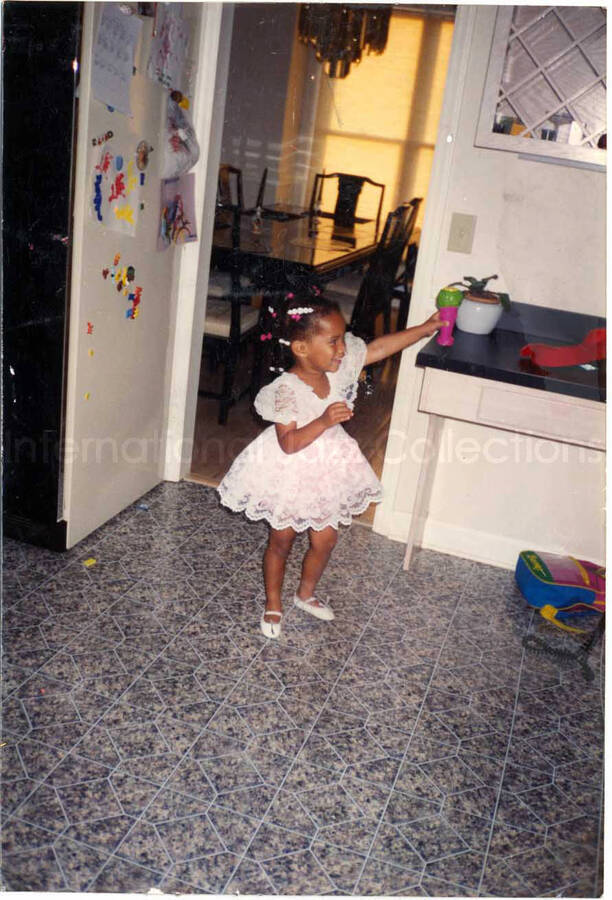 5 x 3 1/2 inch photograph. A girl wearing a lace white dress at home. Handwritten on the back of the photograph: Charise loves music and dance