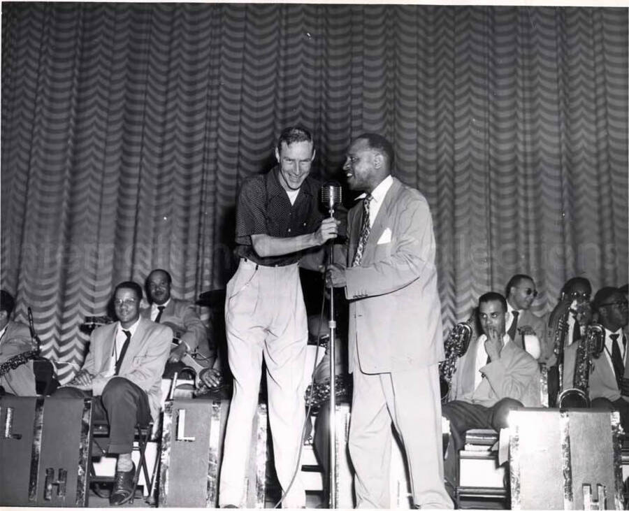 8 x 10 inch photograph. Lionel Hampton on stage with unidentified man. Musicians in the background includes guitarist Billy Mackel
