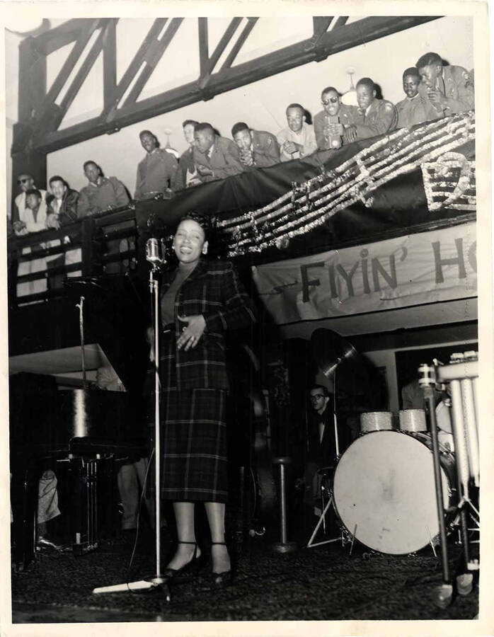 10 x 8 inch photograph. Unidentified vocalist performing for a military audience. A banner in the background reads: Flyin' Home