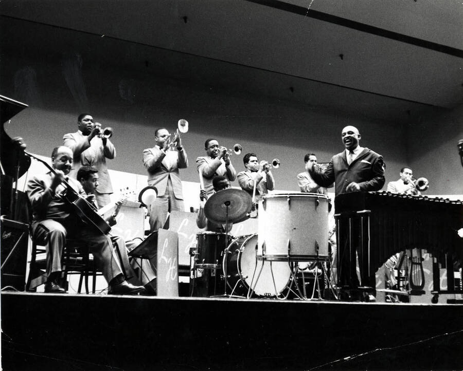 7 1 /2 x 9 1/2 inch photograph. Lionel Hampton with band. William Mackel is on the guitar on the far left. The third trumpeter from the left is Eddie Mullens and Lionel Hampton stands near the vibraphone conducting.