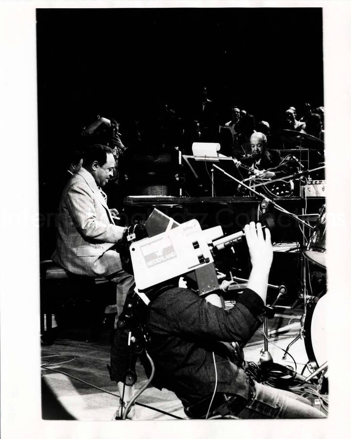 10 x 8 inch photograph. Lionel Hampton playing the piano with orchestra, which includes guitarist Billy Mackel. A cameraman is holding a camera that reads: Nederland [Netherlands?]