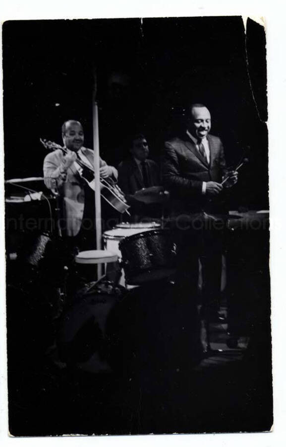 5 1/2 x 3 1/2 inch photograph in the format of postcard. Lionel Hampton on stage with musicians, which includes guitarist Billy Mackel. This postcard was mailed to Lionel Hampton to Royal Orleans Hotel, New Orleans, LA, by photographer Porter. It reads on the back: You can blush, the guitarist can pick; the drummer can beat, but Al hurts. I'll see you tomorrow to finish taking photos. Porter