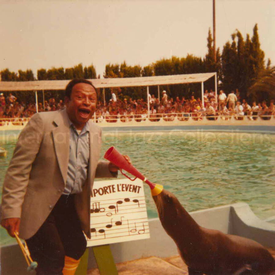 3 1/2 x 3 1/2 inch photograph. Lionel Hampton plays with a sea lion at a marine mammal park. Partially seen above a musical score are the French words: emporte l'event