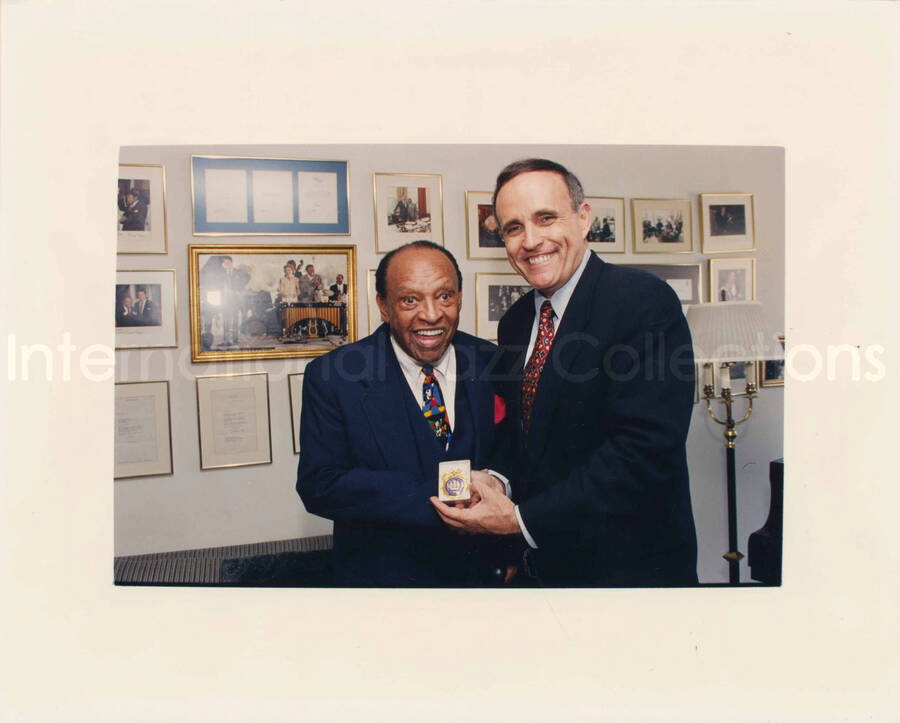 8 x 10 inch photograph. Lionel Hampton with Rudolph Giuliani, Mayor of New York City. They hold a seal of the city pin in a box. They are at Lionel Hampton's apartment, standing in front of a wall displaying plaques and certificates