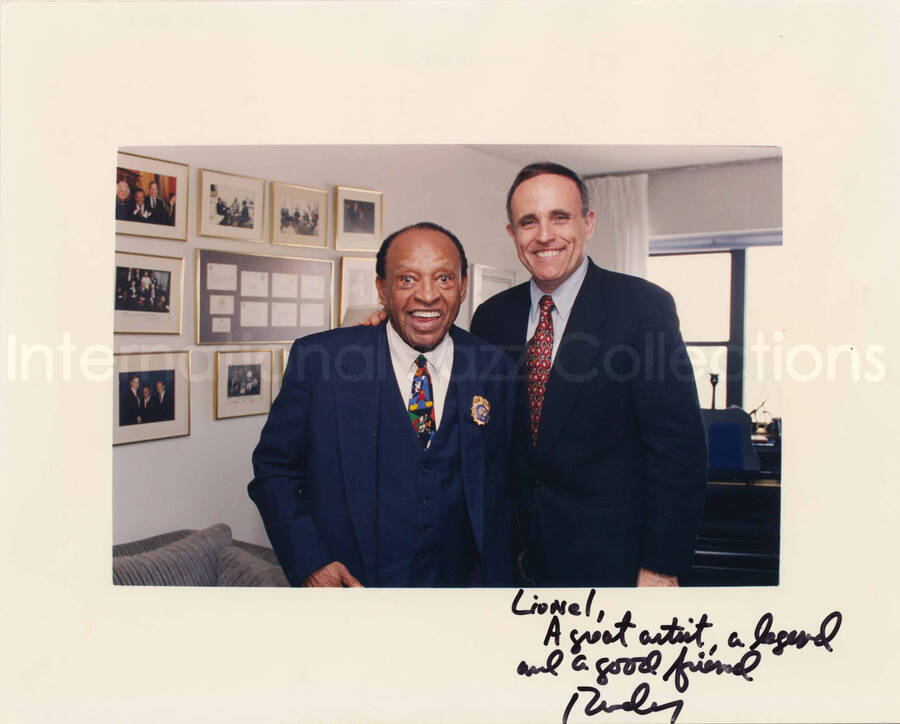 8 x 10 inch signed photograph. Lionel Hampton with Rudolph Giuliani, Mayor of New York City. They are holding a seal of the city pin in a box. They are at Lionel Hampton's apartment, standing in front of a wall displaying plaques and certificates. This photograph is dedicated to Lionel Hampton